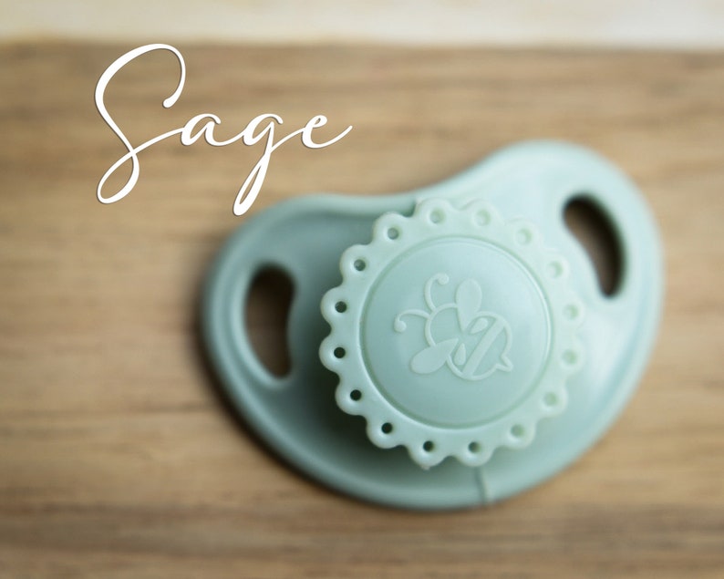 May include: A sage green silicone pacifier with a bee design on the front. The pacifier has a textured, scalloped edge.