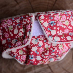 May include: A red cloth diaper with a floral pattern of pink and blue flowers on a white background. The diaper has a white interior and a white Velcro closure. The diaper is designed for babies.