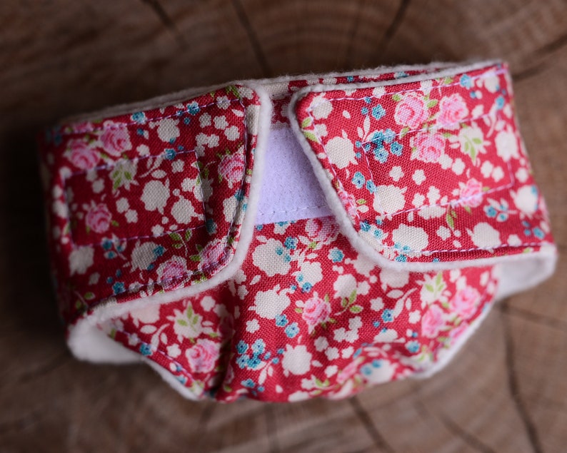 May include: A red cloth diaper with a floral pattern of pink and blue flowers on a white background. The diaper has a white interior and a white Velcro closure. The diaper is designed for babies.
