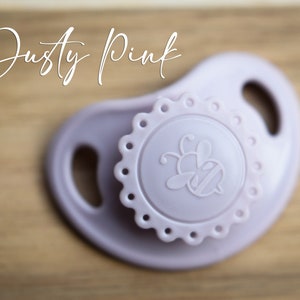 May include: A dusty pink silicone pacifier with a bee design and a scalloped edge.
