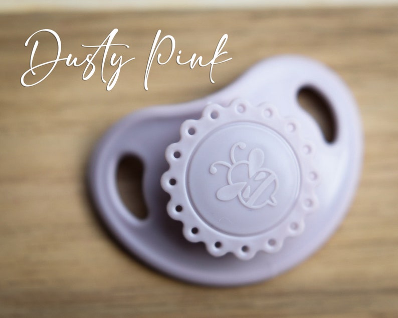 May include: A dusty pink silicone pacifier with a bee design and a scalloped edge.