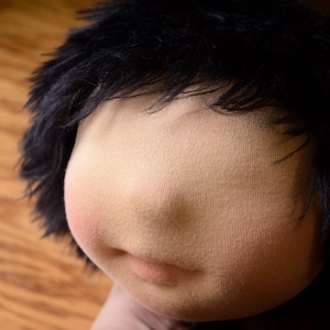May include: A close-up of a doll's face with black hair and a light brown complexion. The doll has a simple, stitched mouth and no visible eyes.