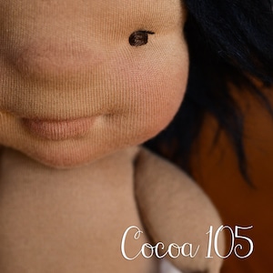 Op de afbeelding: Handgemaakte pop met een bruine huidskleur, zwart haar en een gestikte gezichtsuitdrukking. Het lichaam van de pop is zichtbaar en de tekst "Cocoa 105" staat in cursief op de afbeelding.