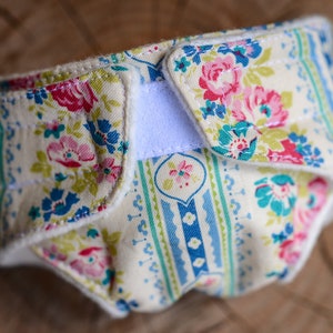 May include: A cloth diaper with a floral pattern in pink, blue, and green on a cream background. The diaper has a white Velcro closure and is made of soft fabric. The design features roses and other flowers.