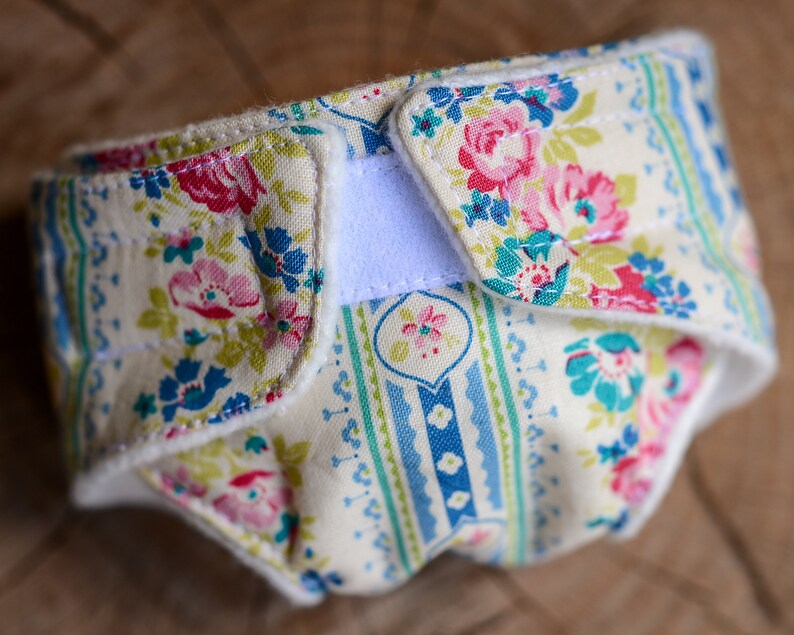 May include: A cloth diaper with a floral pattern in pink, blue, and green on a cream background. The diaper has a white Velcro closure and is made of soft fabric. The design features roses and other flowers.
