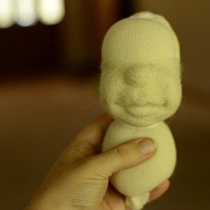 May include: A hand holds a white felt doll with a simple face and a smile. The doll is made from a sock and has a round body.
