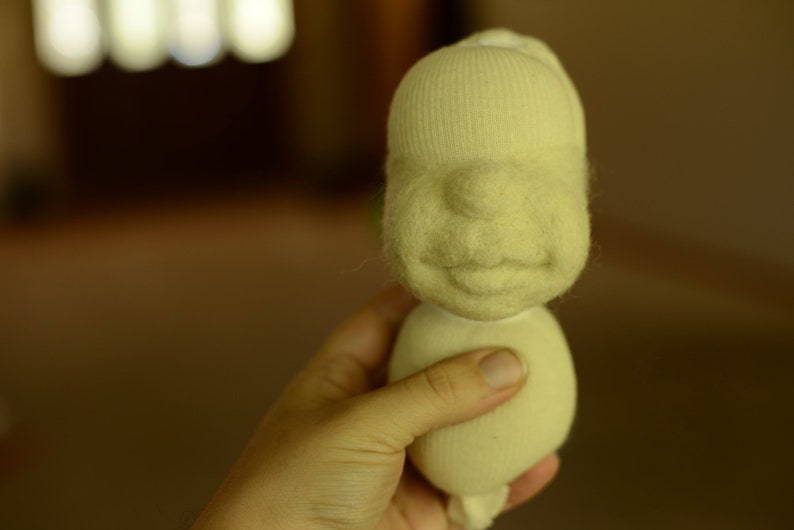 May include: A hand holds a white felt doll with a simple face and a smile. The doll is made from a sock and has a round body.