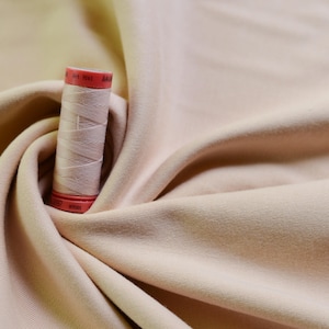 Puede incluir: Primer plano de una tela beige con un carrete de hilo beige en la parte superior. El carrete de hilo tiene etiquetas rojas y blancas con el texto "Art. 1061" y "100m".