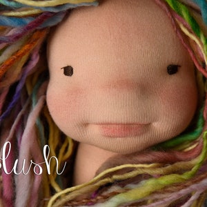 Op de afbeelding: Een handgemaakte poppenkop met een zacht, stoffen gezicht en veelkleurig garen haar. De pop heeft roze wangen, kleine zwarte ogen en een delicate roze mond. Het woord "Blush" staat in cursiefschrift linksonder.