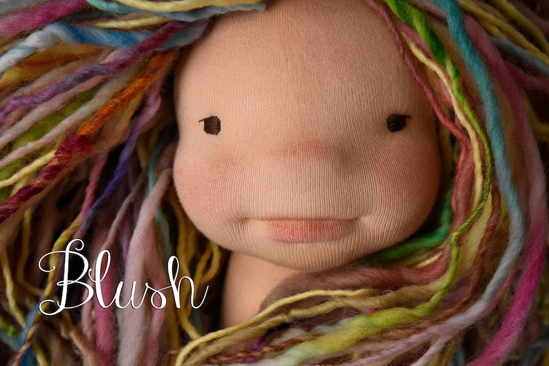 Op de afbeelding: Een handgemaakte poppenkop met een zacht, stoffen gezicht en veelkleurig garen haar. De pop heeft roze wangen, kleine zwarte ogen en een delicate roze mond. Het woord "Blush" staat in cursiefschrift linksonder.