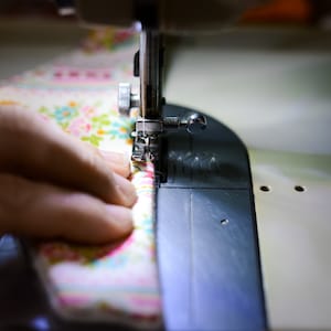 May include: Close-up of a sewing machine in operation, stitching floral fabric. The fabric features a pattern of pink, blue, and yellow flowers on a cream background. The needle is piercing the fabric, guided by a hand. The machine is silver and black.