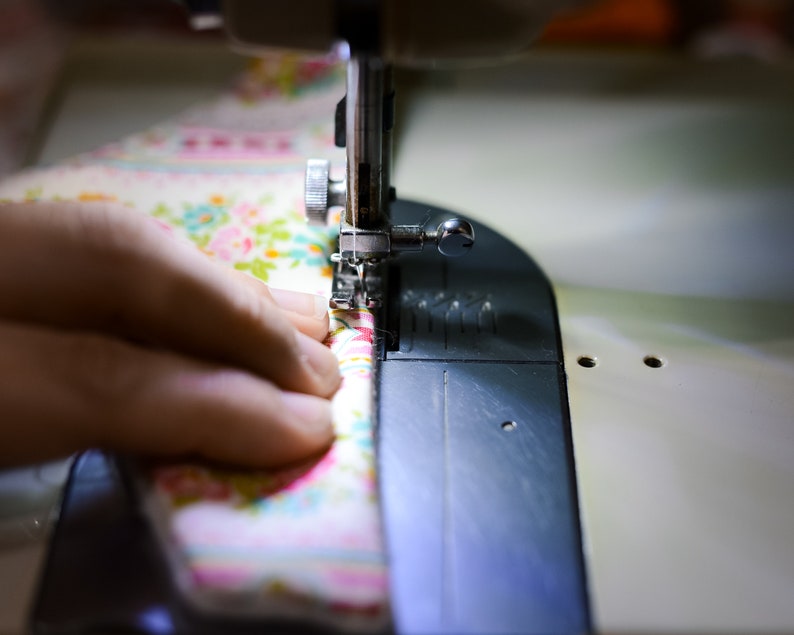 May include: Close-up of a sewing machine in operation, stitching floral fabric. The fabric features a pattern of pink, blue, and yellow flowers on a cream background. The needle is piercing the fabric, guided by a hand. The machine is silver and black.