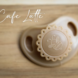 May include: A beige pacifier with a bee design on the front. The pacifier has a scalloped edge and is made of silicone.