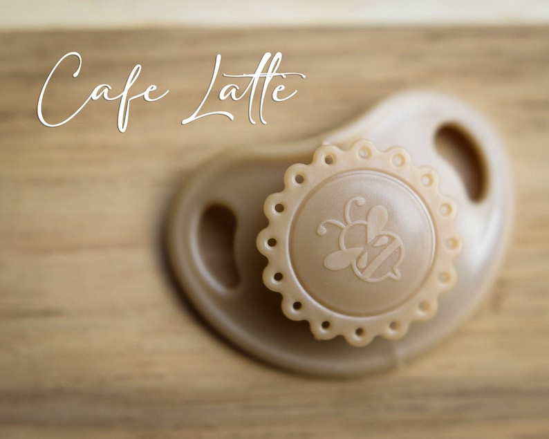 May include: A beige pacifier with a bee design on the front. The pacifier has a scalloped edge and is made of silicone.