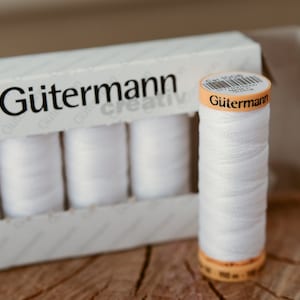 May include: A box of white Gutermann Creative thread spools with a single spool of white thread sitting in front of the box. The spool has a yellow label with the text "Gutermann" on it.
