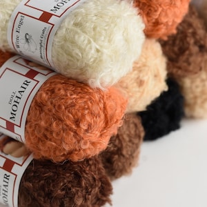 K&ouml;nnte beinhalten: Eine Sammlung von lockigen Mohairgarnstr&auml;ngen in verschiedenen Schattierungen von Creme, Orange, Braun und Schwarz. Jeder Strang ist mit einer wei&szlig;en Papierbanderole umwickelt, die den Text Doll Mohair und Witte Engel sowie ein Engel-Symbol zeigt.