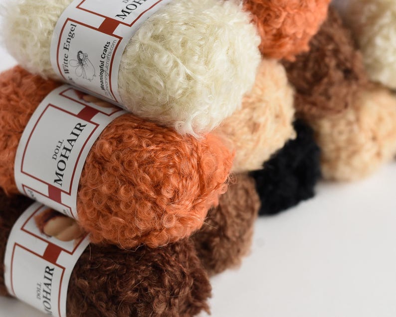 K&ouml;nnte beinhalten: Eine Sammlung von lockigen Mohairgarnstr&auml;ngen in verschiedenen Schattierungen von Creme, Orange, Braun und Schwarz. Jeder Strang ist mit einer wei&szlig;en Papierbanderole umwickelt, die den Text Doll Mohair und Witte Engel sowie ein Engel-Symbol zeigt.