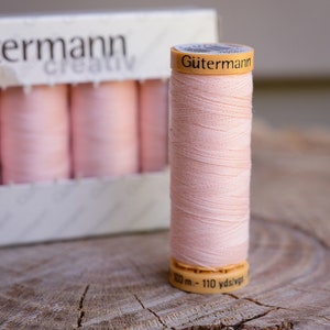 May include: A spool of pale pink Gütermann thread with a yellow label that reads "Gütermann" and "100 m - 110 yds/vgs".