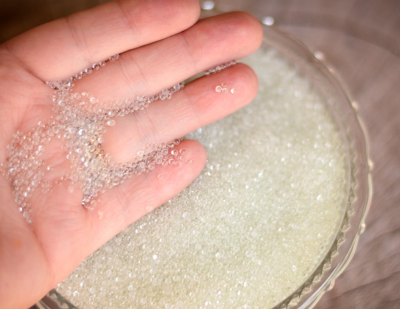 May include: A hand holding a handful of clear glass beads, with a bowl of the same beads in the background.