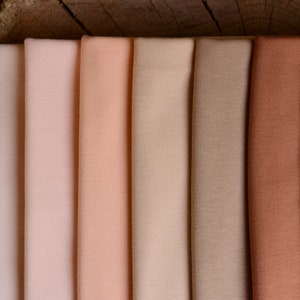 Op de afbeelding: Stalen van stoffen in een reeks neutrale kleuren, van lichtbeige tot diep bruin. De stof is netjes opgevouwen en toont de verschillende tinten en texturen. De achtergrond is een houten oppervlak.
