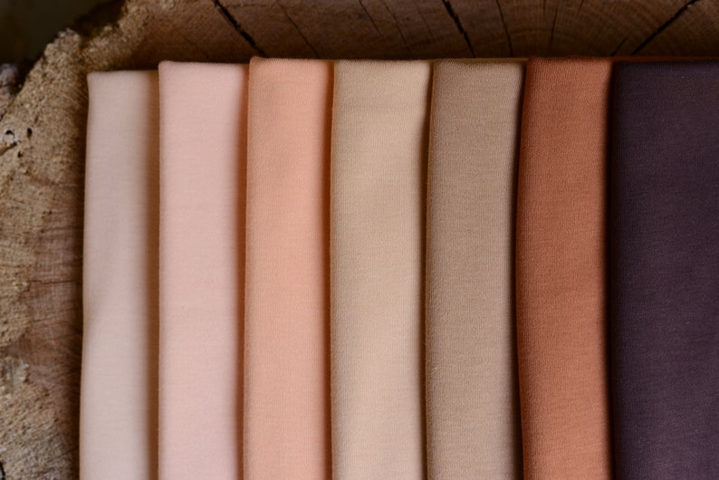 Op de afbeelding: Stalen van stoffen in een reeks neutrale kleuren, van lichtbeige tot diep bruin. De stof is netjes opgevouwen en toont de verschillende tinten en texturen. De achtergrond is een houten oppervlak.
