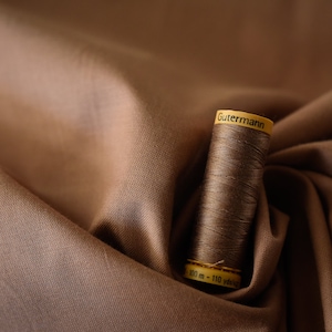 Könnte beinhalten: Eine Spule braunen Fadens mit einem gelben Etikett, auf dem "Gutermann" und "100 m - 110 yds" steht, auf einem braunen Stoffhintergrund.