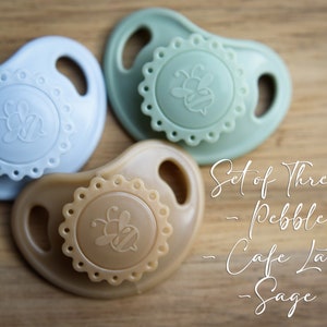 May include: Set of three silicone baby pacifiers in pebble, cafe latte, and sage colors. Each pacifier features a bee design and a scalloped edge.