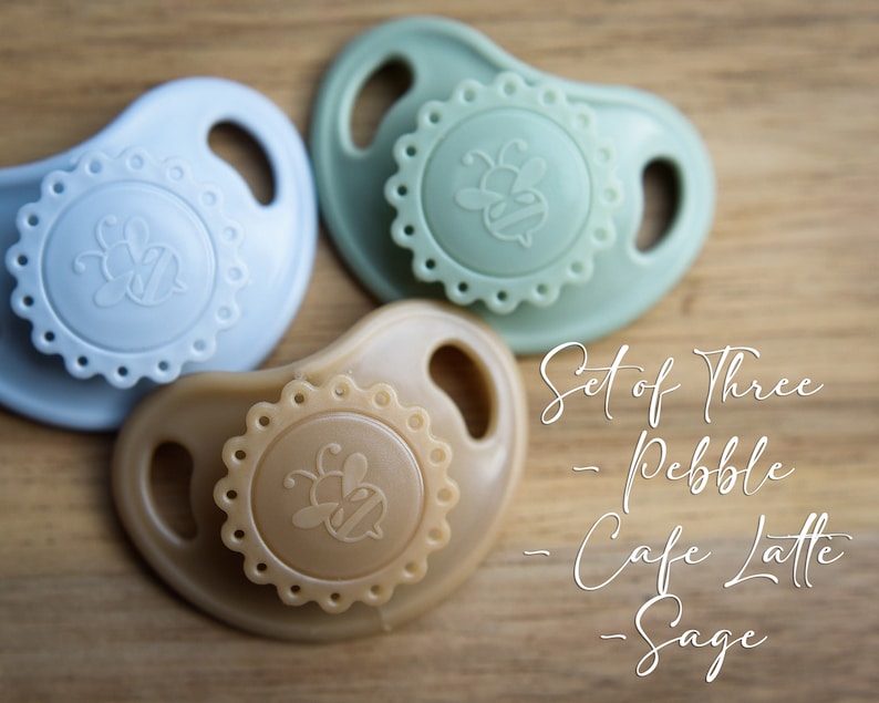 May include: Set of three silicone baby pacifiers in pebble, cafe latte, and sage colors. Each pacifier features a bee design and a scalloped edge.