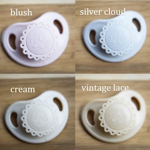 May include: Four pacifiers in different colors: blush pink, silver cloud, cream, and vintage lace. Each pacifier has a decorative design with a bee in the center.