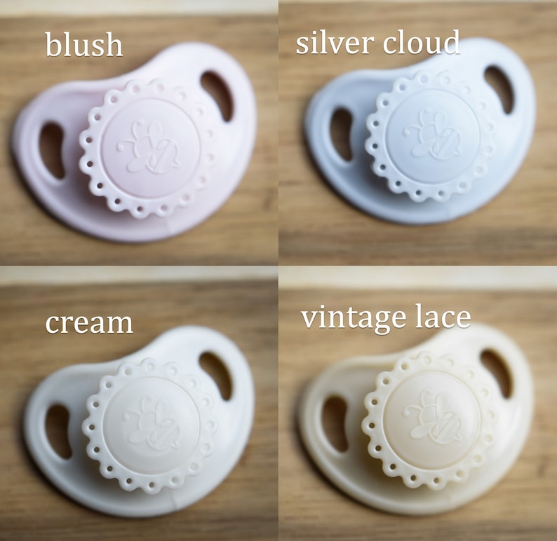 May include: Four pacifiers in different colors: blush pink, silver cloud, cream, and vintage lace. Each pacifier has a decorative design with a bee in the center.