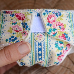 May include: A floral patterned cloth diaper with a cream background, featuring pink, blue, and green flowers. The diaper has a white velcro closure and is held in a hand. The fabric has a quilted appearance.