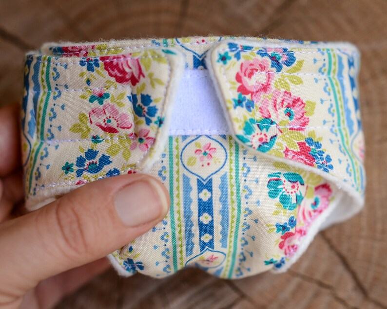 May include: A floral patterned cloth diaper with a cream background, featuring pink, blue, and green flowers. The diaper has a white velcro closure and is held in a hand. The fabric has a quilted appearance.