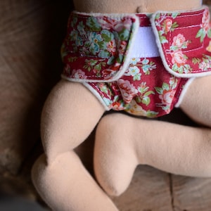 May include: A doll wearing a red cloth diaper with a floral pattern of pink and blue flowers. The diaper has a white closure and trim. The doll's legs are visible, made of a soft, beige fabric.