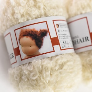 K&ouml;nnte beinhalten: Ein Kn&auml;uel cremefarbenes, lockiges Mohairgarn mit einem Papieretikett, das den Kopf einer Puppe mit roten Haaren zeigt. Das Etikett enth&auml;lt die Angaben 64 % Mohair, 27 % Wolle und 9 % Polyamid bei einem Gewicht von ca. 50 Gramm pro Kn&auml;uel.