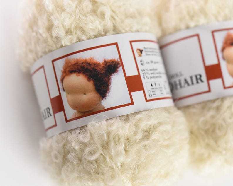 K&ouml;nnte beinhalten: Ein Kn&auml;uel cremefarbenes, lockiges Mohairgarn mit einem Papieretikett, das den Kopf einer Puppe mit roten Haaren zeigt. Das Etikett enth&auml;lt die Angaben 64 % Mohair, 27 % Wolle und 9 % Polyamid bei einem Gewicht von ca. 50 Gramm pro Kn&auml;uel.