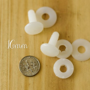 K&ouml;nnte beinhalten: Vier wei&szlig;e Kunststoff-Sicherheitsaugen mit einem Durchmesser von 1 cm. Jedes Auge hat eine Schraubstift und eine Unterlegscheibe. Die Augen befinden sich auf einer Holzoberfl&auml;che.