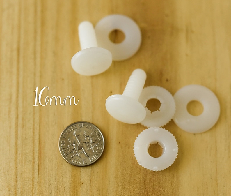 K&ouml;nnte beinhalten: Vier wei&szlig;e Kunststoff-Sicherheitsaugen mit einem Durchmesser von 1 cm. Jedes Auge hat eine Schraubstift und eine Unterlegscheibe. Die Augen befinden sich auf einer Holzoberfl&auml;che.