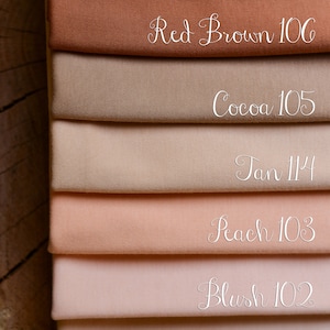 Op de afbeelding: Een stapel stofstalen in bruin- en roze tinten. De stalen zijn gelabeld met kleurnamen en nummers, waaronder "Dark Brown 104", "Red Brown 106", "Cocoa 105", "Tan 114", "Peach 103", "Blush 102" en "Fair 115".