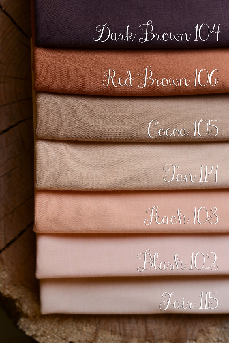 Op de afbeelding: Een stapel stofstalen in bruin- en roze tinten. De stalen zijn gelabeld met kleurnamen en nummers, waaronder "Dark Brown 104", "Red Brown 106", "Cocoa 105", "Tan 114", "Peach 103", "Blush 102" en "Fair 115".