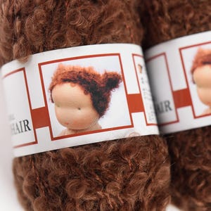 K&ouml;nnte beinhalten: Zwei Kn&auml;uel lockiges braunes Garn mit einem wei&szlig;en Papieretikett, das eine Grafik eines Puppenkopfes mit passendem braunem Haar zeigt. Das Etikett enth&auml;lt den Text "DOLL HAIR" in schwarzer Schrift und einen roten Rahmen um die Illustration.