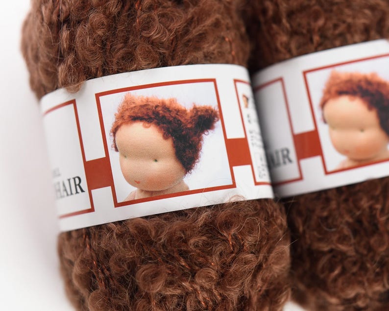 K&ouml;nnte beinhalten: Zwei Kn&auml;uel lockiges braunes Garn mit einem wei&szlig;en Papieretikett, das eine Grafik eines Puppenkopfes mit passendem braunem Haar zeigt. Das Etikett enth&auml;lt den Text "DOLL HAIR" in schwarzer Schrift und einen roten Rahmen um die Illustration.