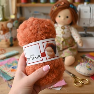 K&ouml;nnte beinhalten: Ein Kn&auml;uel aus lockigem, rostorangem Mohairgarn mit einem wei&szlig;en Papieretikett, das den Text DOLL MOHAIR und eine Abbildung eines Puppenkopfes zeigt. Das Garn hat eine flauschige, strukturierte Optik und wird vor einer Stoffpuppe mit rotem Haar gehalten.