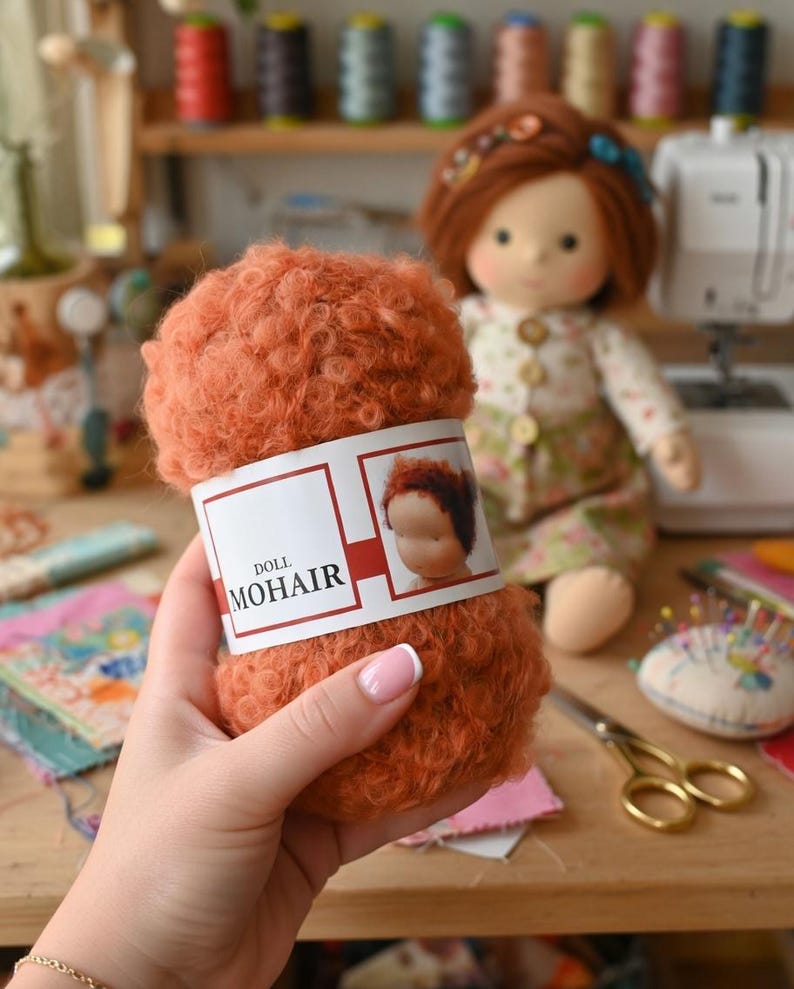 K&ouml;nnte beinhalten: Ein Kn&auml;uel aus lockigem, rostorangem Mohairgarn mit einem wei&szlig;en Papieretikett, das den Text DOLL MOHAIR und eine Abbildung eines Puppenkopfes zeigt. Das Garn hat eine flauschige, strukturierte Optik und wird vor einer Stoffpuppe mit rotem Haar gehalten.