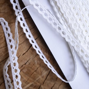 May include: White crochet lace trim with a scalloped edge and small circles. The lace is rolled up on a white surface.