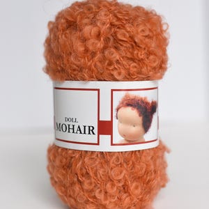K&ouml;nnte beinhalten: Ein Kn&auml;uel aus lockigem, leuchtend orangefarbenem Mohairgarn f&uuml;r die Puppenherstellung. Das mittlere Papieretikett zeigt den Text DOLL MOHAIR und eine kleine Abbildung eines Puppenkopfes mit passendem orangefarbenem Haar. Das Garn ist strukturiert.