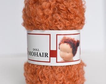 May include: A ball of curly, vibrant orange mohair yarn for doll making. The center paper label features the text DOLL MOHAIR and a small illustration of a doll head with matching orange hair. The yarn has a distinct, textured, curly appearance.
