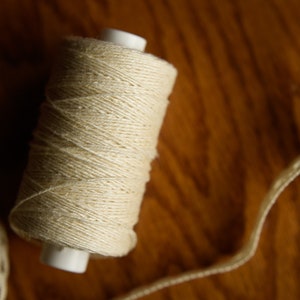 Puede incluir: Una bobina de hilo de algod&oacute;n blanco natural con una bobina de pl&aacute;stico blanca. El hilo est&aacute; enrollado apretado y tiene una textura lisa.