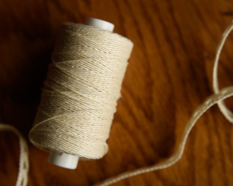 Puede incluir: Una bobina de hilo de algod&oacute;n blanco natural con una bobina de pl&aacute;stico blanca. El hilo est&aacute; enrollado apretado y tiene una textura lisa.