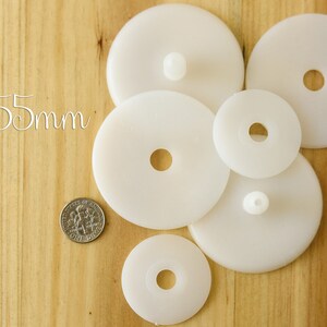 K&ouml;nnte beinhalten: F&uuml;nf wei&szlig;e Plastikkreise mit einem Loch in der Mitte. Die Kreise sind unterschiedlich gro&szlig;, der gr&ouml;&szlig;te Kreis hat einen Durchmesser von 55 mm. Die Kreise sind zuf&auml;llig auf einer Holzoberfl&auml;che angeordnet.
