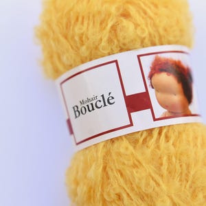 K&ouml;nnte beinhalten: Ein Kn&auml;uel aus leuchtend gelbem Mohair-Boucl&eacute;-Garn mit strukturierter, lockiger Optik. Ein wei&szlig;es Papieretikett umschlie&szlig;t die Mitte und zeigt den Text Mohair Boucl&eacute; sowie eine kleine Abbildung eines Puppenkopfes mit rotbraunem Haar.